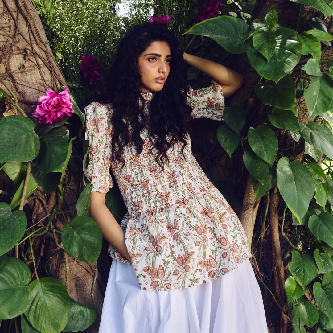Woman in a floral dress standing among greenery with pink flowers.
