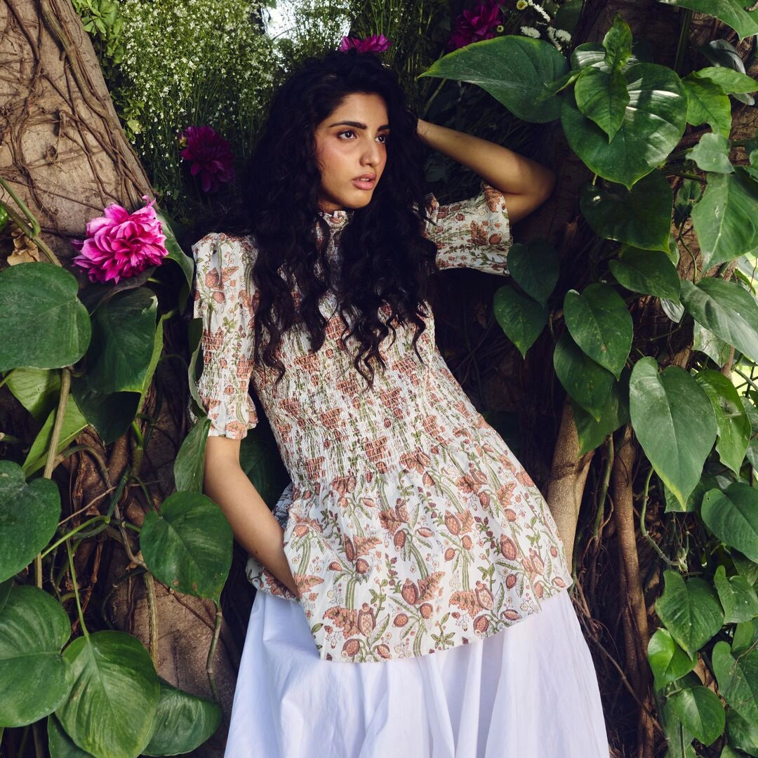 Woman in a floral dress standing among greenery with pink flowers.