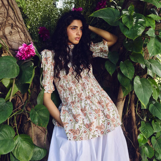 Woman in a floral dress standing among greenery with pink flowers.