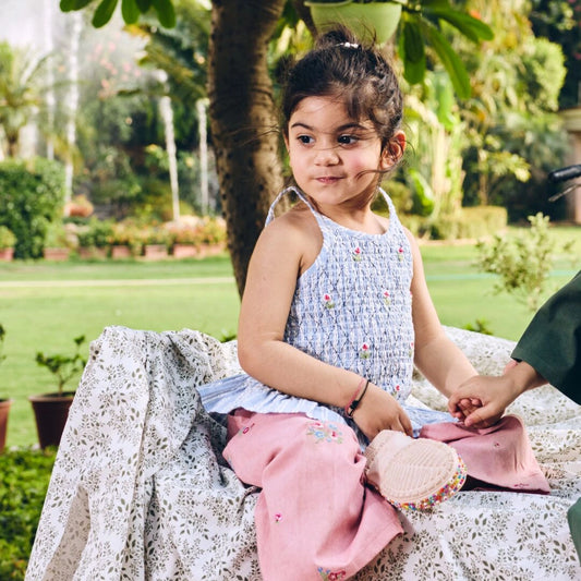 Young girl sitting outdoors holding a pink bag, with greenery in the background