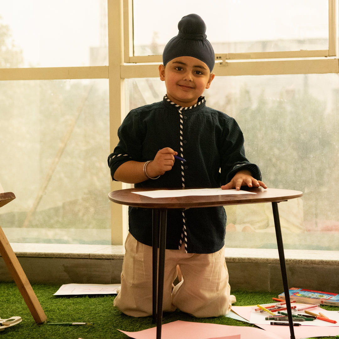 Child in traditional attire standing behind a small table with art supplies on a grass-like surface.