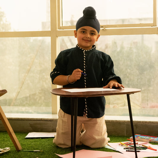 Child in traditional attire standing behind a small table with art supplies on a grass-like surface.