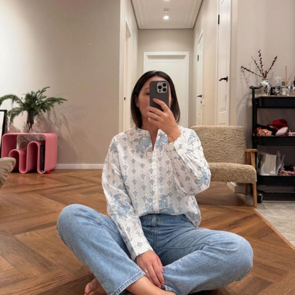 Person sitting on the floor taking a mirror selfie wearing a floral blouse and jeans.