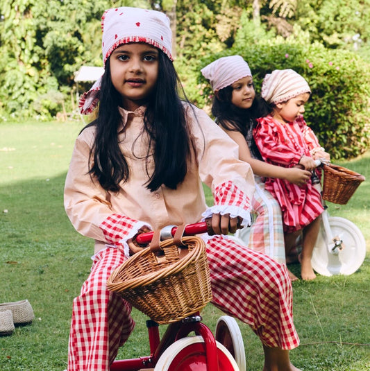 Three young girls in matching outfits with checkered dresses and headbands riding tricycles in a park.
