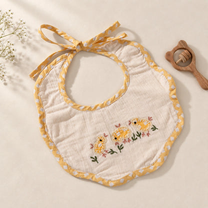A baby bib made of soft white muslin fabric with yellow polka-dot trim and hand embroidery embroidered yellow chicks with pink flowers on the front. The bib has a tie-neck design.