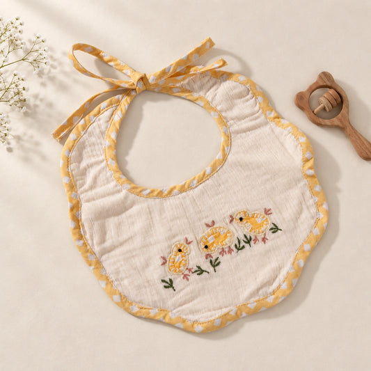 A baby bib made of soft white muslin fabric with yellow polka-dot trim and hand embroidery embroidered yellow chicks with pink flowers on the front. The bib has a tie-neck design.