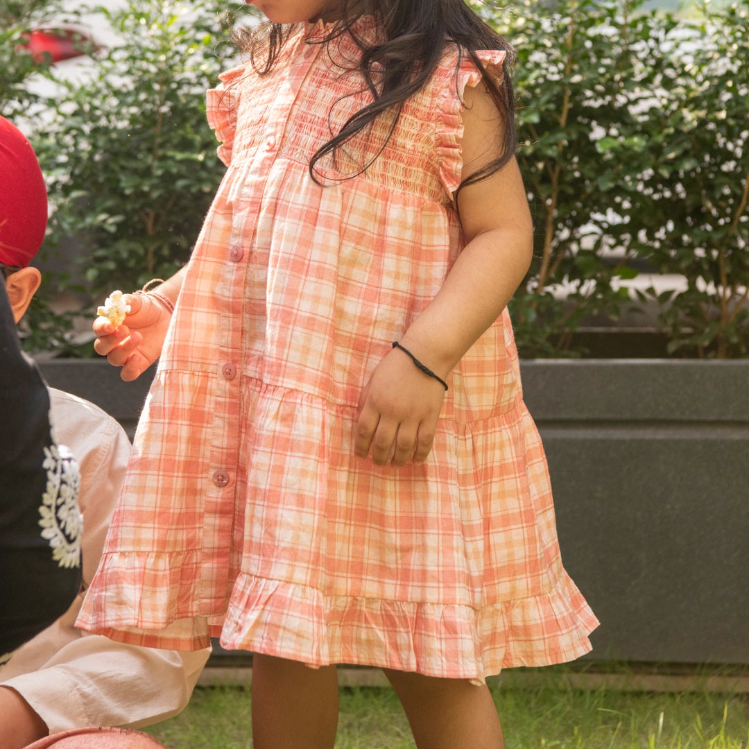 A child wearing a sleeveless peach and white plaid dress with ruffle details.