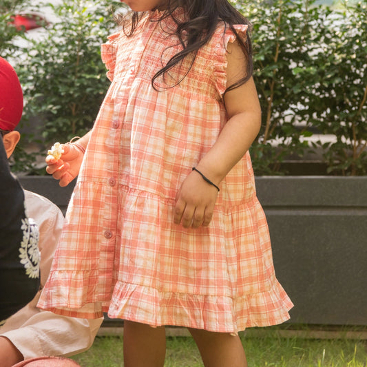 A child wearing a sleeveless peach and white plaid dress with ruffle details.