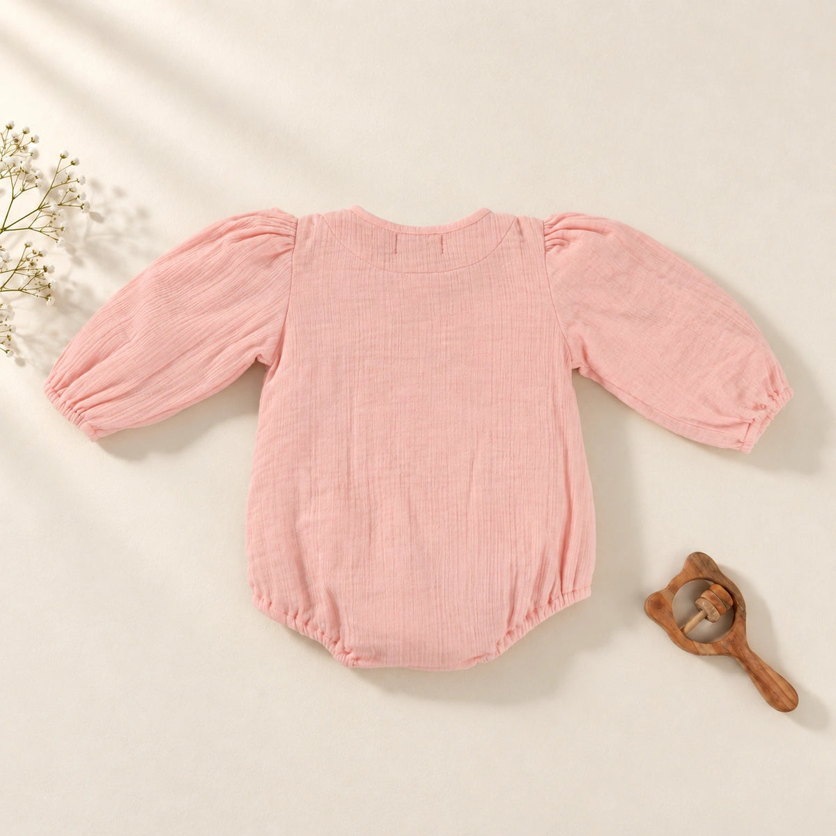 A baby long-sleeve romper in textured pink muslin fabric, featuring a ruffled collar with two front buttons and elasticized cuffs and leg openings. 