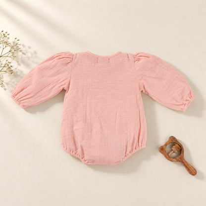 A baby long-sleeve romper in textured pink muslin fabric, featuring a ruffled collar with two front buttons and elasticized cuffs and leg openings. 