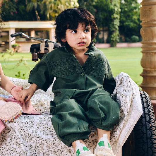 Child in a green outfit sitting on a textured surface with a garden background