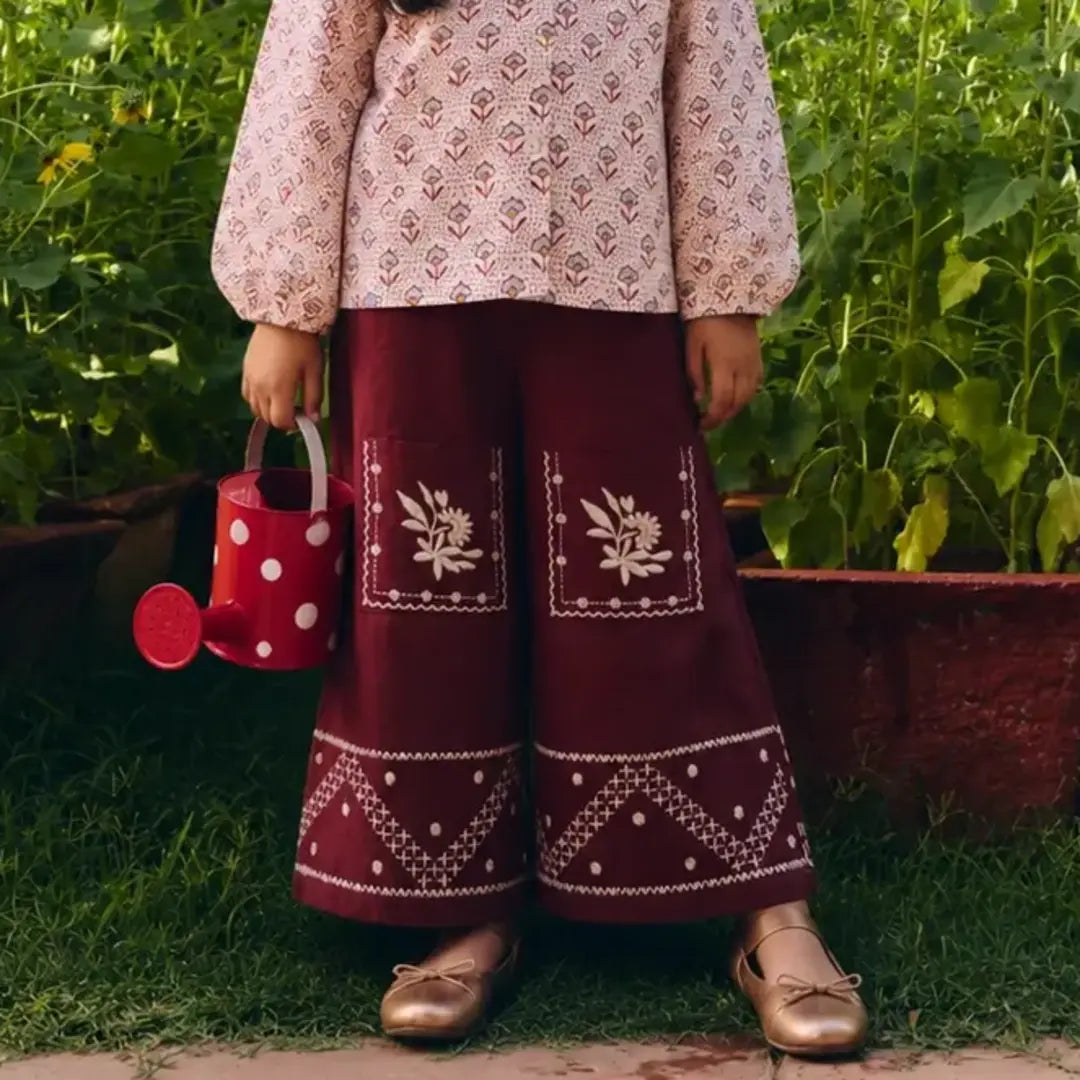 A child standing outdoors in a garden wearing  wide-leg burgundy pants with white embroidered floral and geometric details. 