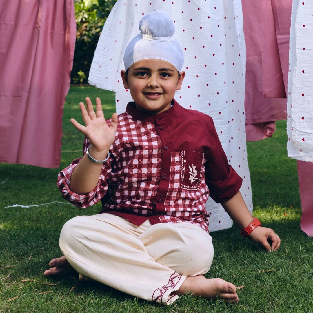 A young boy sitting on green grass outdoors, wearing a traditional outfit with a maroon and white gingham kurta, cream trousers, and a white skullcap. Pink and white patterned garments are hanging in the background.