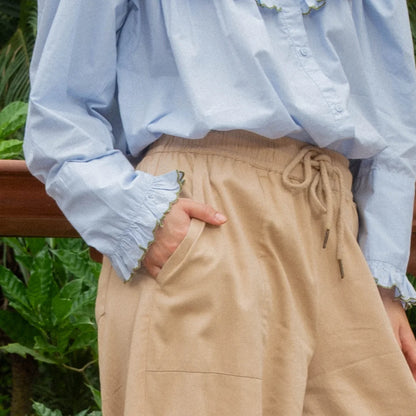A close-up of a person wearing beige drawstring wide-leg pants and a light blue long-sleeve blouse, standing outdoors near green foliage.
