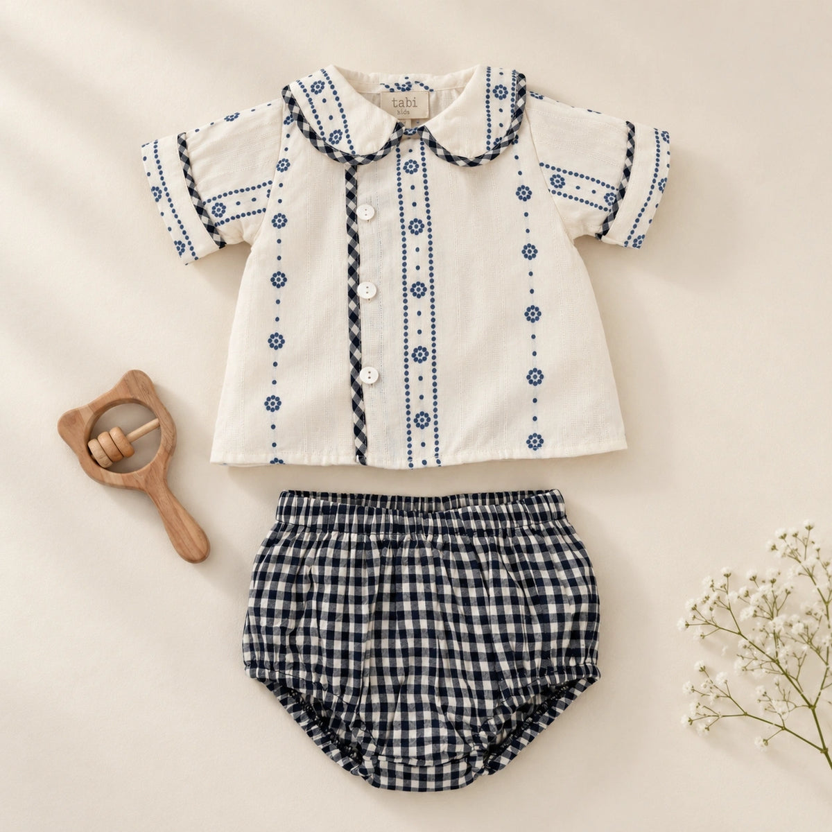 A baby clothing set  featuring a short-sleeve cream shirt with blue embroidered patterns and a black and white gingham bloomer. 