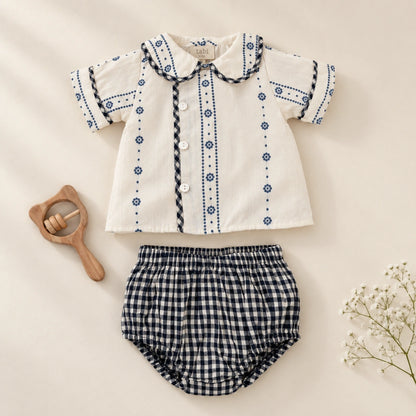 A baby clothing set  featuring a short-sleeve cream shirt with blue embroidered patterns and a black and white gingham bloomer. 