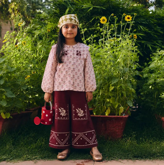 A young girl stands outdoors in a garden wearing patterned long-sleeve top and deep maroon wide-leg pants with  embroidery. 