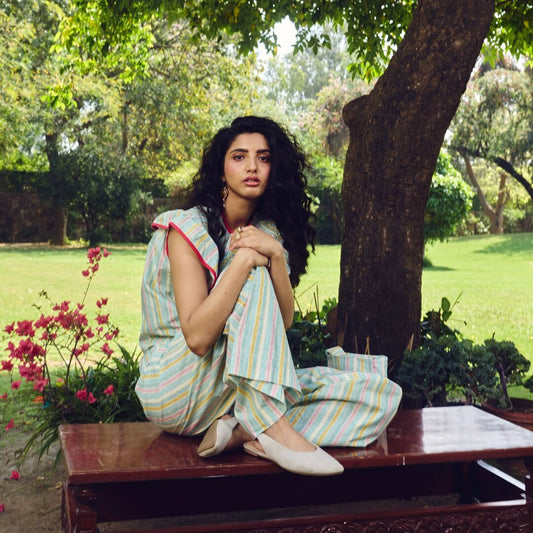 Woman in a striped outfit sitting on a bench in a park with trees and flowers around.