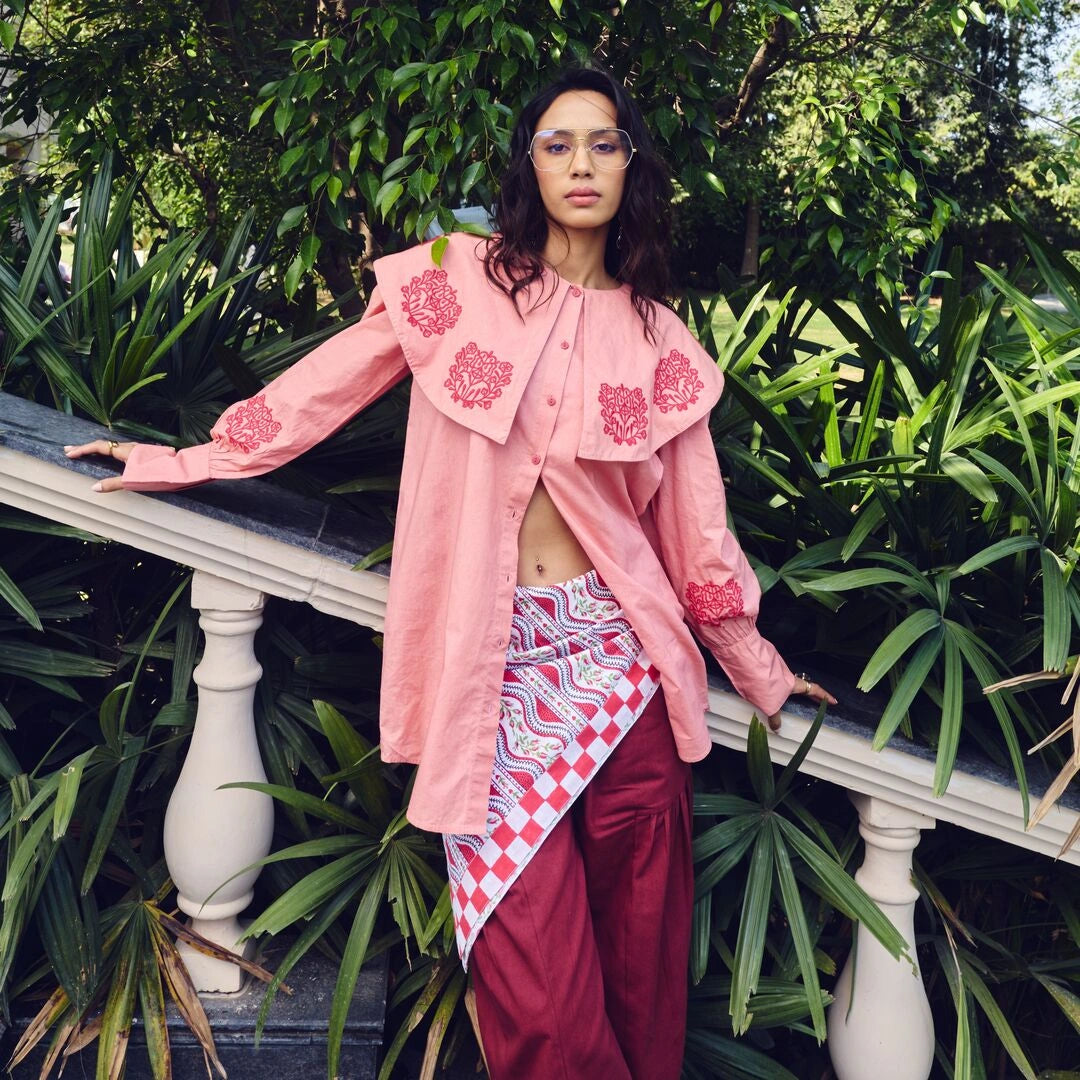Person wearing a pink embroidered shirt and red pants standing outdoors with greenery.