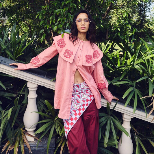 Person wearing a pink embroidered shirt and red pants standing outdoors with greenery.