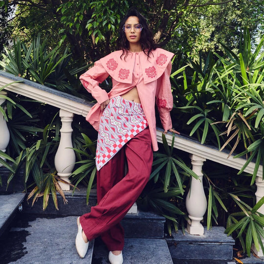 Person wearing a pink floral blouse and red pants standing outdoors with greenery.