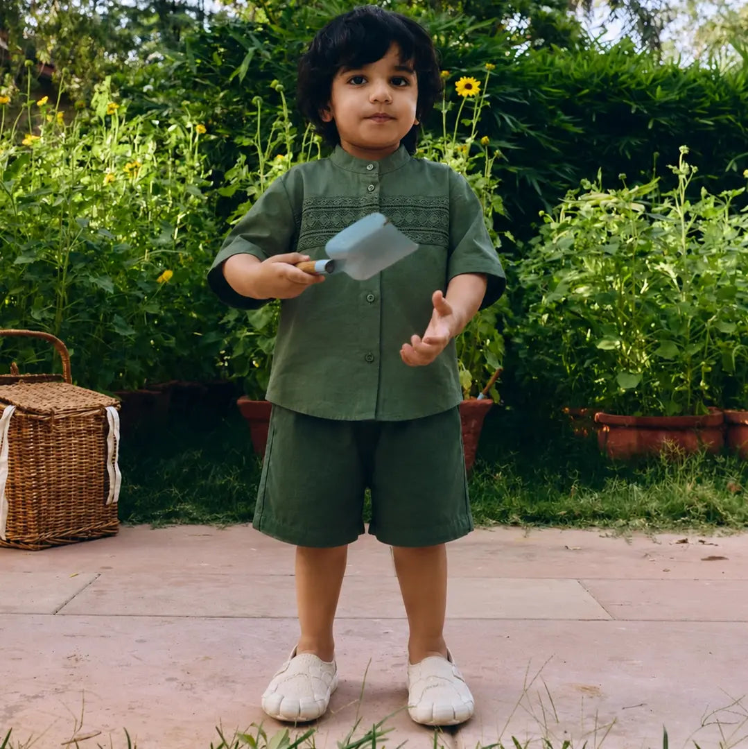 A young child wearing an olive green short-sleeve button-up shirt and matching shorts.