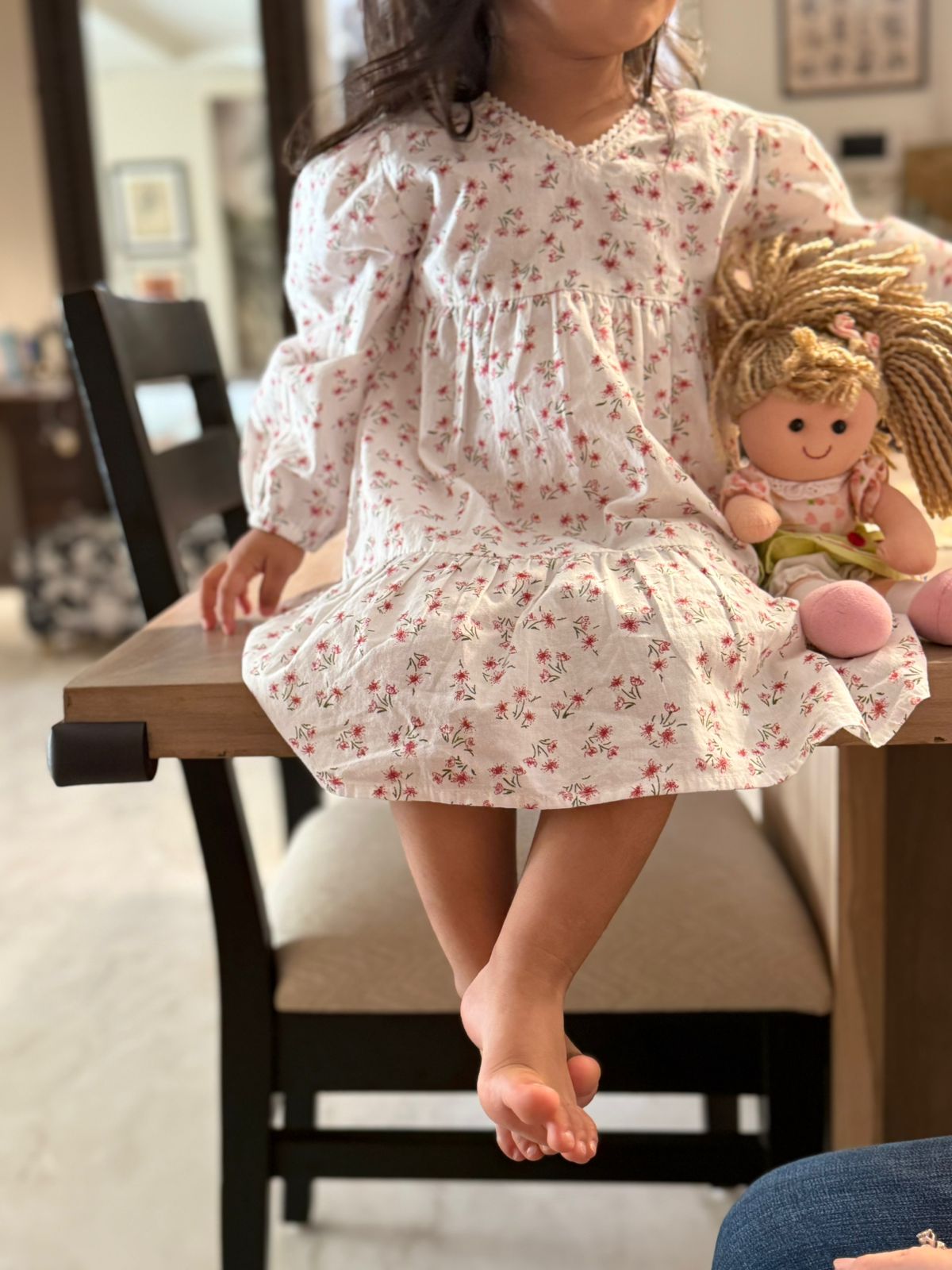 Child in a floral dress holding a doll, sitting on a chair indoors.