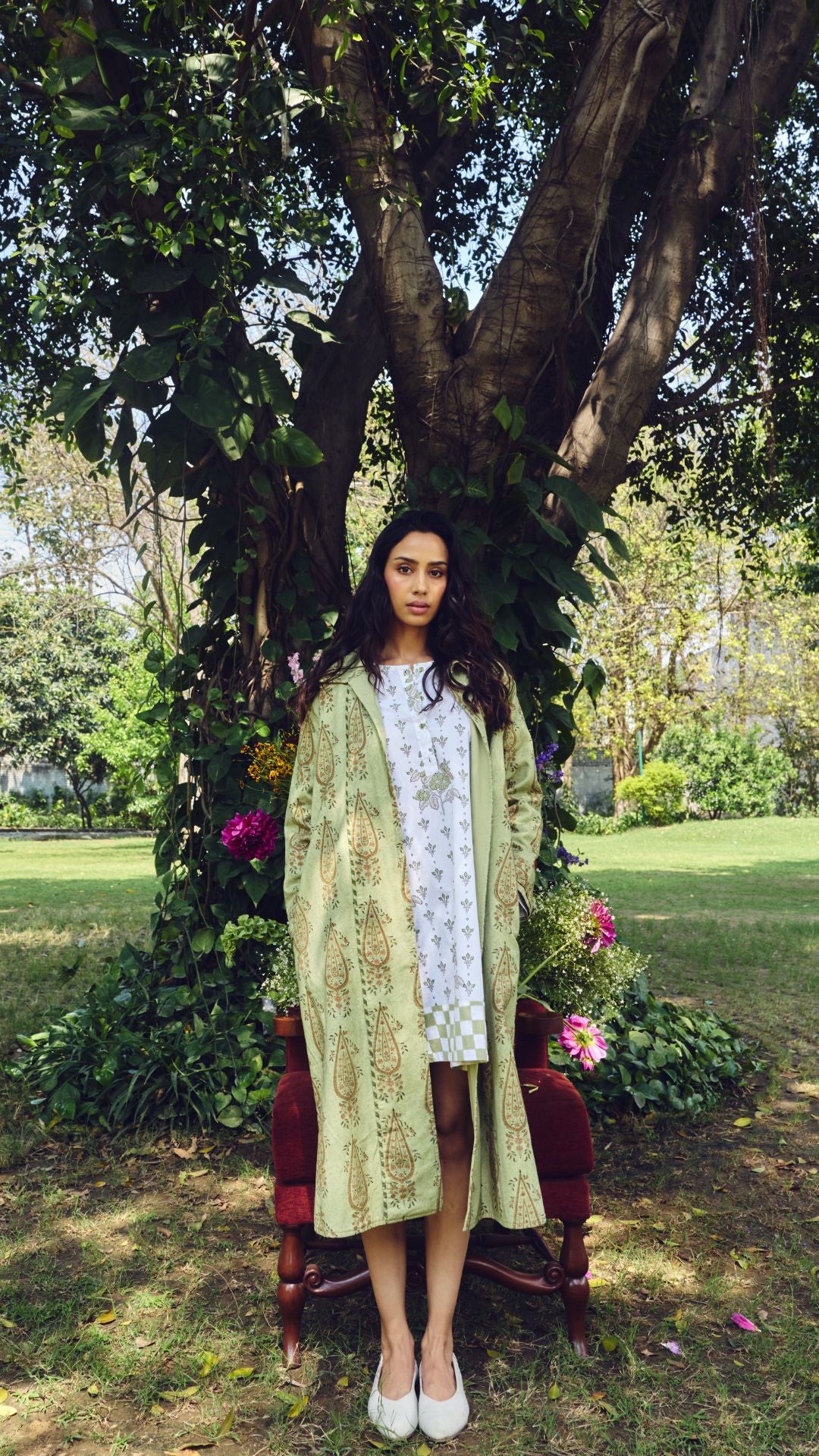 A woman  wearing a long light green patterned jacket over a white printed dress.
