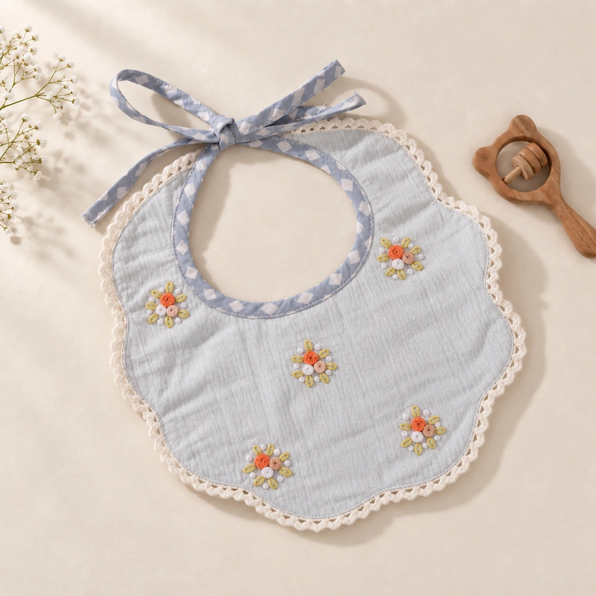 A blue baby bib featuring delicate floral hand embroidery, adjustable tie-up closures and contrasting checked piping.