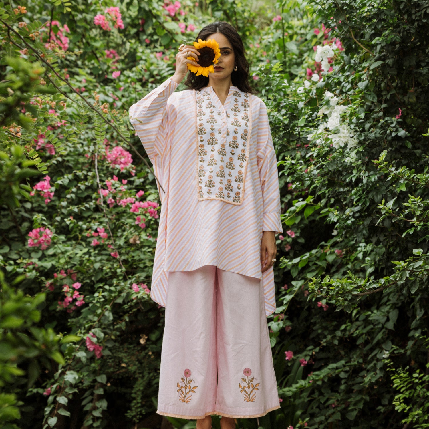 Woman in a light pink traditional outfit with floral embroidery standing in a garden.