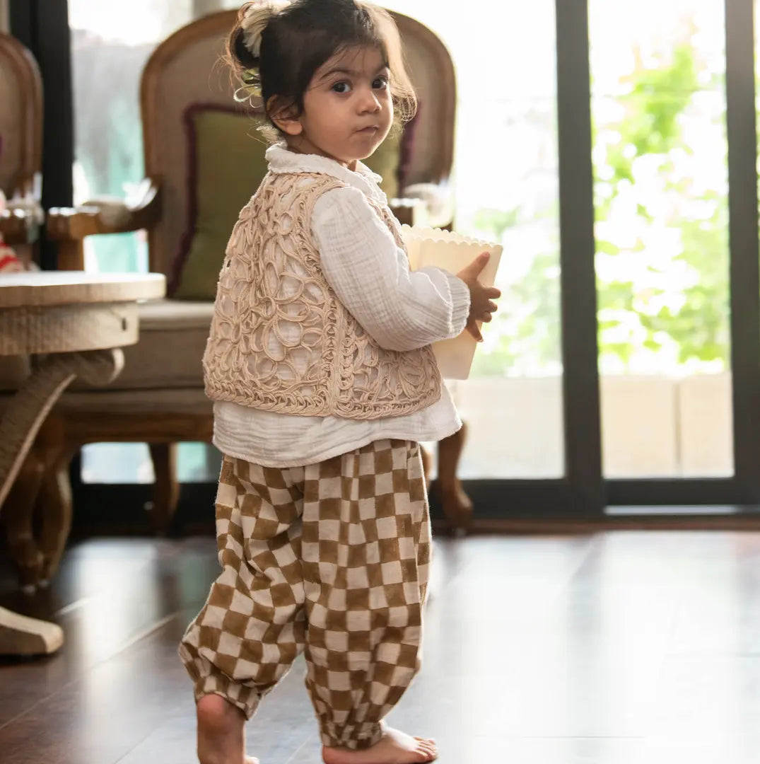 A toddler girl wearing a cream lace vest over a white long-sleeve top and tan-and-cream checkered pants.