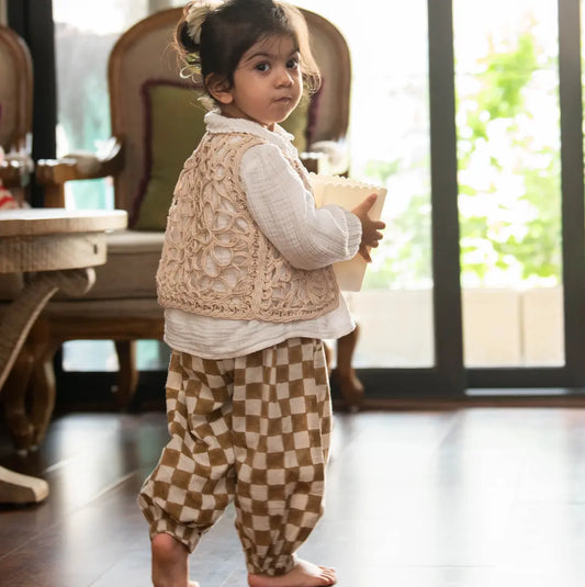 A toddler girl wearing a cream lace vest over a white long-sleeve top and tan-and-cream checkered pants.