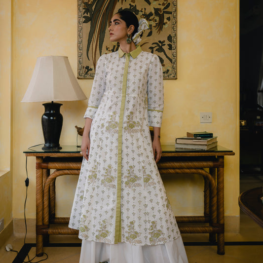 Woman in a white and green embroidered traditional outfit standing in a room with a lamp and books on a table.