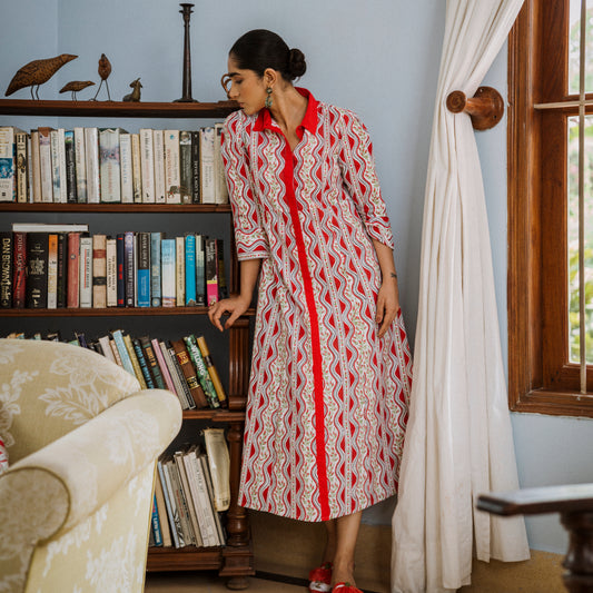 Woman in a red and white patterned dress standing in a room with a bookshelf and window.