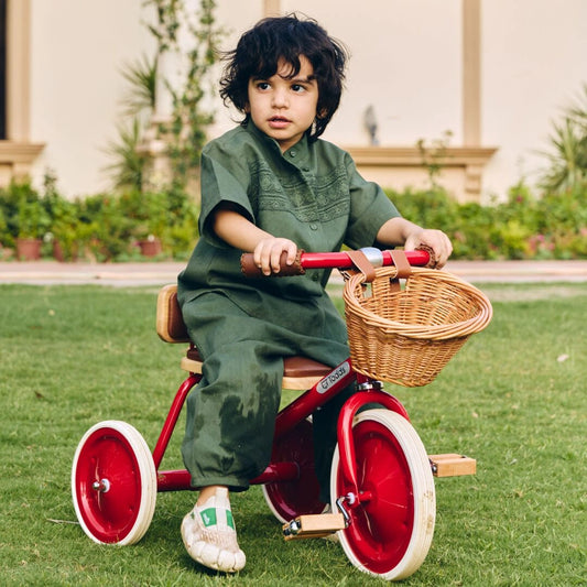 Child in green outfit riding a red tricycle with a basket in an outdoor setting