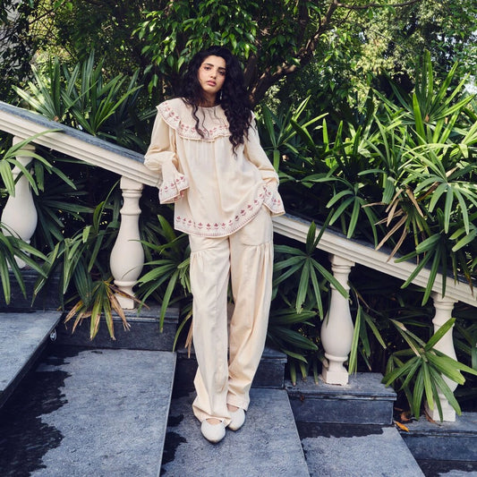 A woman stands on outdoor stone steps surrounded by lush green plants, wearing a cream-colored two-piece lounge set with delicate pink embroidery and ruffled details.