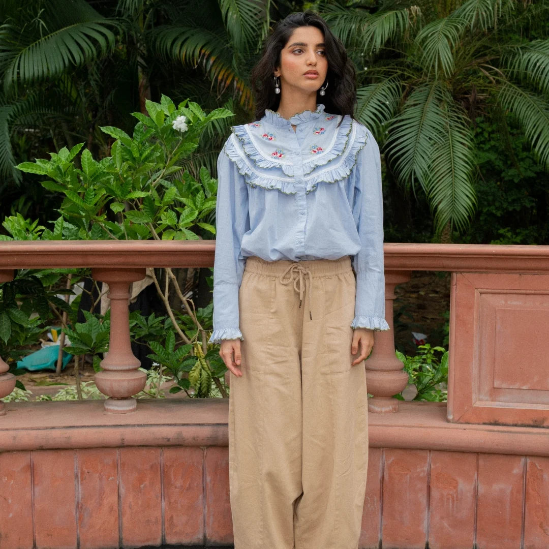 A woman stands outdoors  wearing a light blue long-sleeve blouse with a large ruffled collar and embroidered details, paired with beige wide-leg drawstring pants.