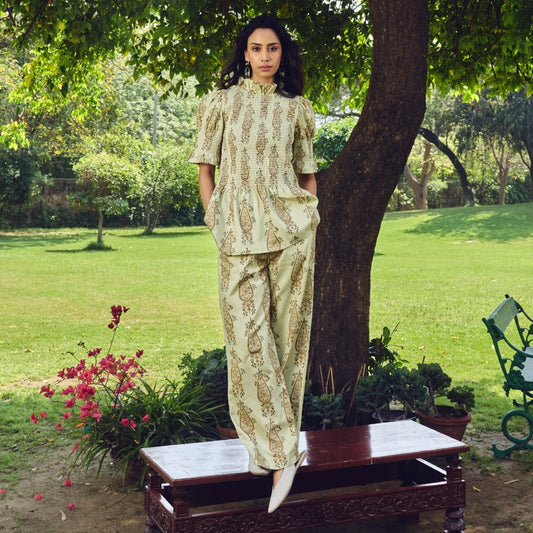 A woman stands outdoors on a bench in a park setting, wearing a matching green and gold patterned two-piece outfit with a high-neck top and wide-leg pants.