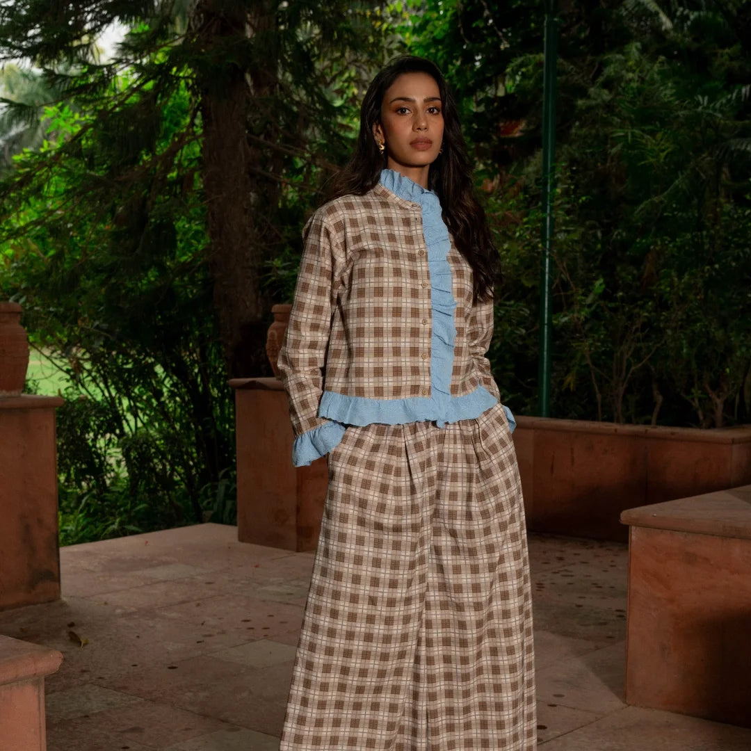 A woman stands outdoors  wearing a long-sleeve beige and brown plaid Top & Pants with a light blue ruffled collar, waist trim, and cuffs. 