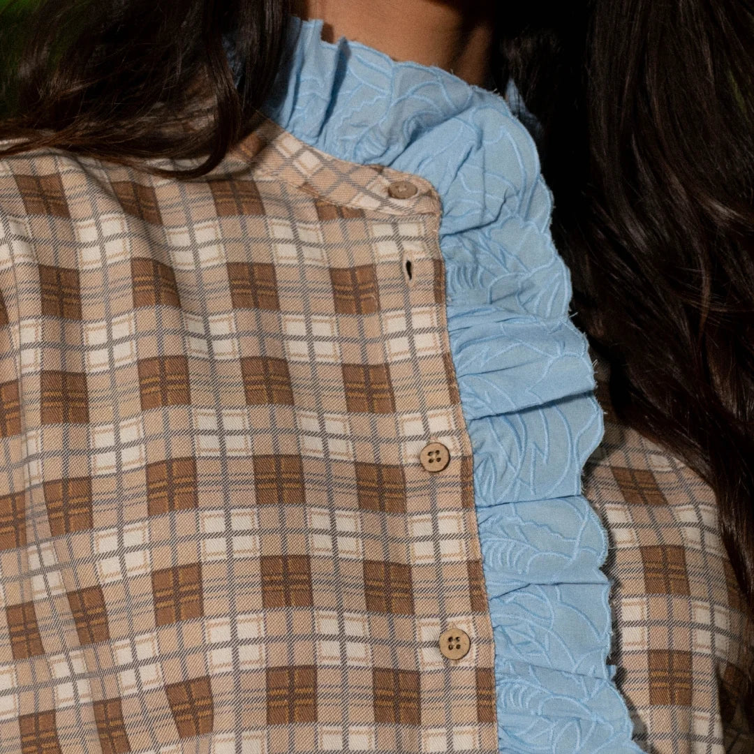 Close-up of a person wearing a brown and beige plaid button-front top layered over a light blue ruffled blouse.