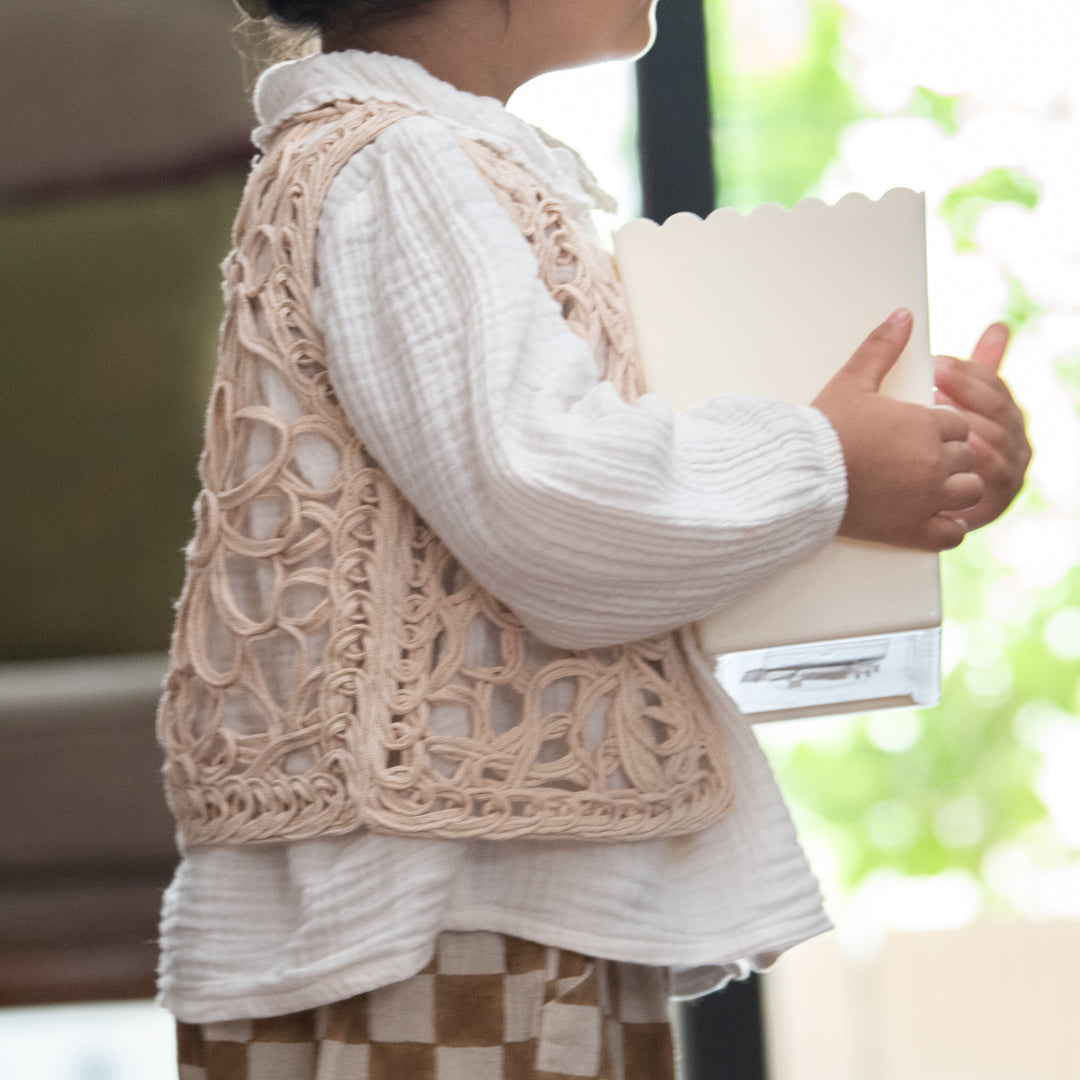 Person wearing a beige lace vest over a white shirt, holding a book.