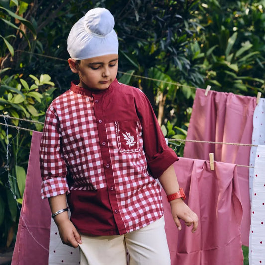 Child wearing a red checkered shirt and white turban standing near a clothesline with pink fabric.