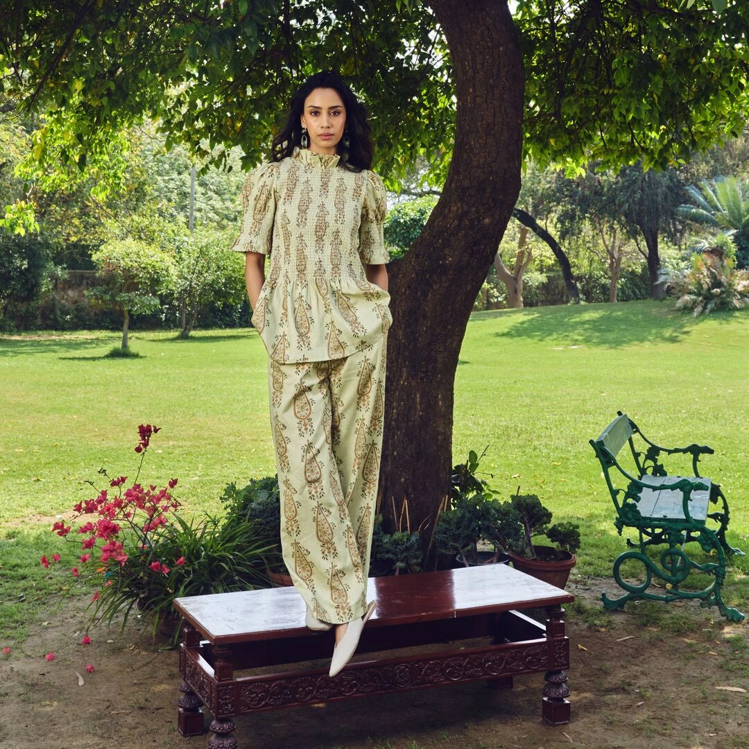 A woman stands outdoors in a park-like garden setting under a large tree, wearing a matching beige and gold patterned short-sleeve top and wide-leg pants. She is posed on a carved wooden bench with a green garden bench and lawn visible in the background.
