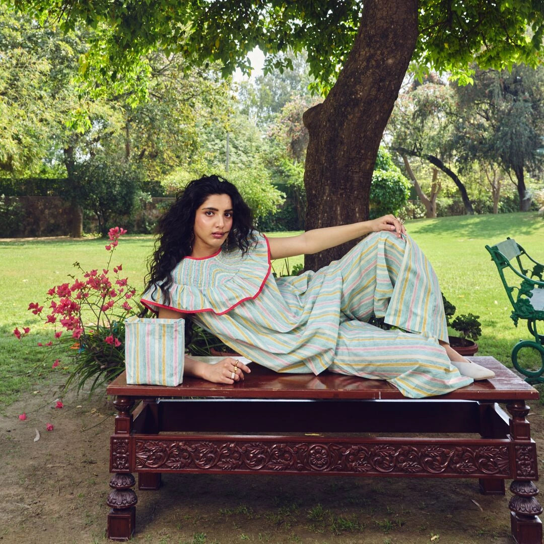 Woman in a striped dress sitting on a wooden bench in a park-like setting with trees and flowers.