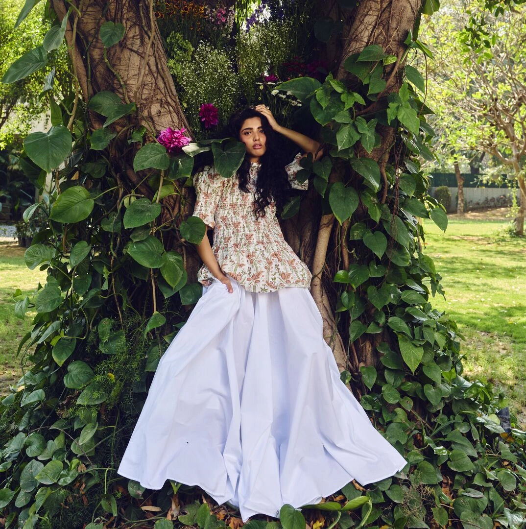 Woman in a floral top and white pants standing in a garden with greenery and flowers.