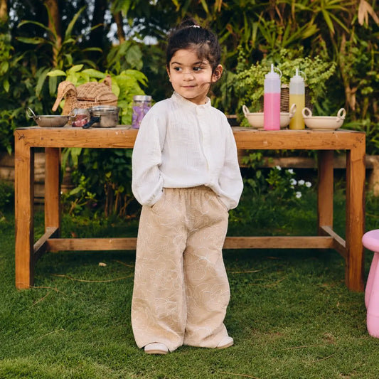 A young child wearing a white long-sleeve top and beige patterned wide-leg pants. 