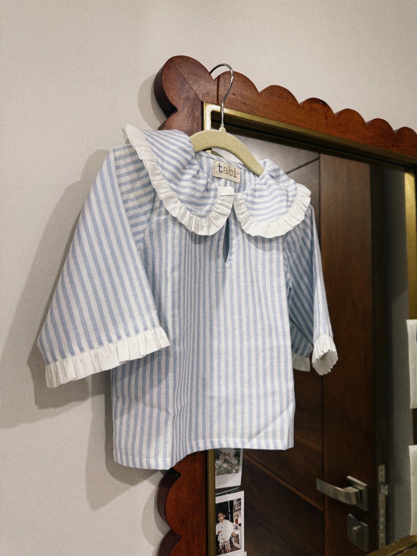 Striped shirt with ruffled collar on a wooden hanger against a neutral wall.