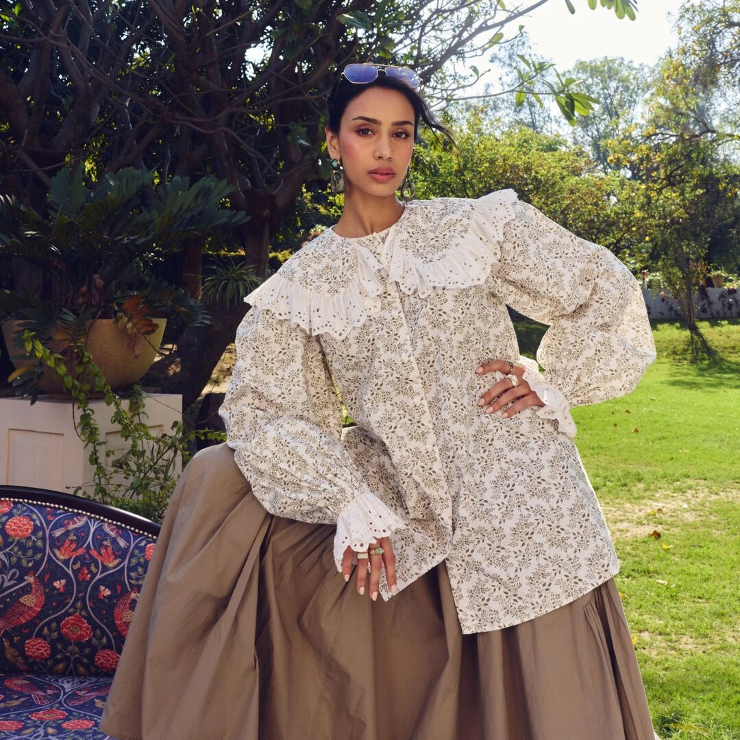 A woman stands outdoors in a garden wearing a cream and white floral print blouse with dramatic ruffled shoulders and a brown pleated skirt. Trees, grass, and patio furniture are visible in the background.