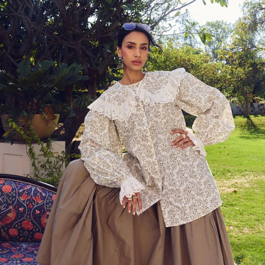 A woman stands outdoors in a garden wearing a cream and white floral print blouse with dramatic ruffled shoulders and a brown pleated skirt. Trees, grass, and patio furniture are visible in the background.