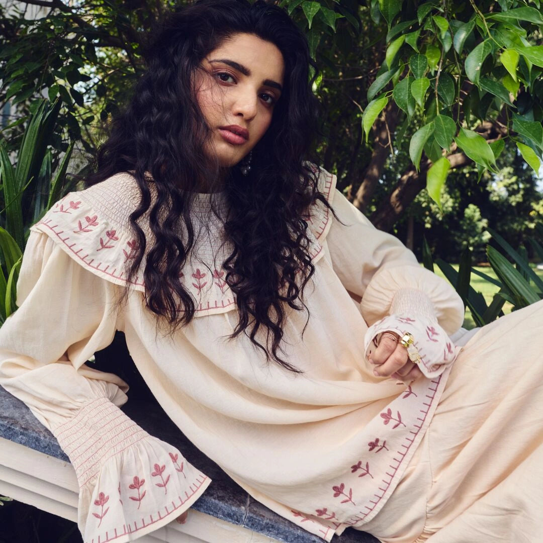 Woman wearing a cream-colored traditional outfit with floral embroidery, sitting outdoors among greenery.